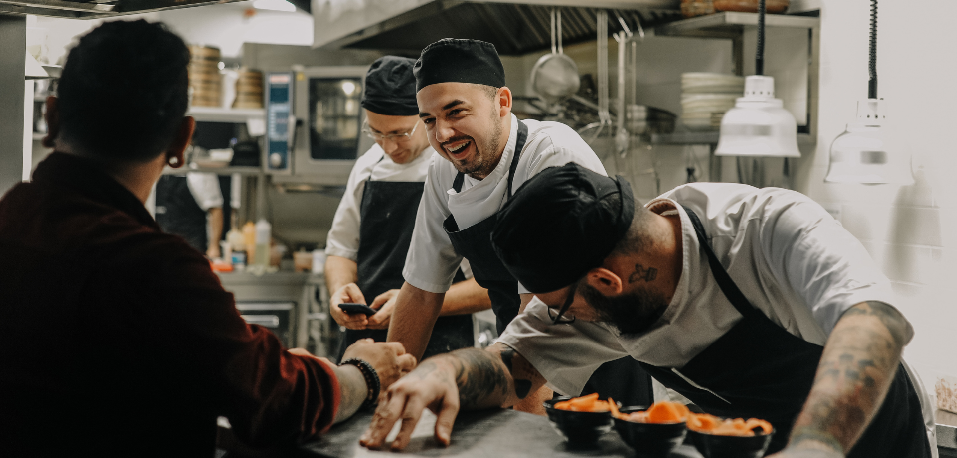Four chefs laughing and talking in a professional kitchen, one leaning on a counter near bowls of sliced carrots.