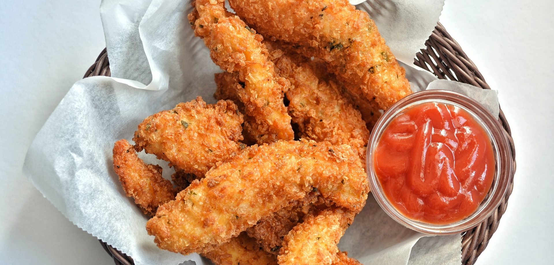 Crispy breaded fried chicken tenders in a basket lined with parchment paper, served with a side of ketchup.