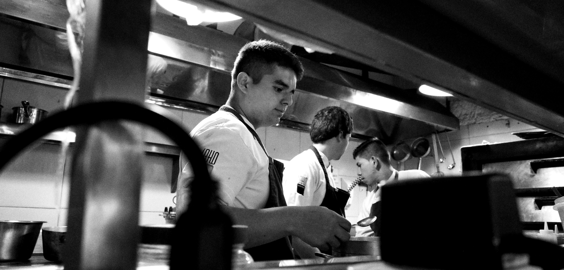 A black and white, wide-angle photograph capturing three kitchen staff members working intently along a line under bright heat lamps.